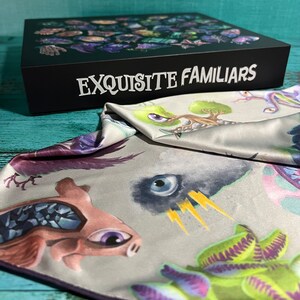 Puede incluir: Una bolsa de tela gris con ribete morado. La bolsa est&aacute; decorada con ilustraciones coloridas de criaturas fant&aacute;sticas, incluyendo una criatura rosa con un ojo grande, una planta verde con zarcillos morados y una criatura negra con un solo ojo. La bolsa est&aacute; doblada sobre una caja negra con el texto "EXQUISITE FAMILIARS" impreso en blanco.