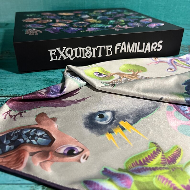 Puede incluir: Una bolsa de tela gris con ribete morado. La bolsa est&aacute; decorada con ilustraciones coloridas de criaturas fant&aacute;sticas, incluyendo una criatura rosa con un ojo grande, una planta verde con zarcillos morados y una criatura negra con un solo ojo. La bolsa est&aacute; doblada sobre una caja negra con el texto "EXQUISITE FAMILIARS" impreso en blanco.