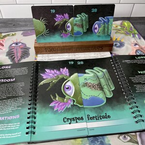 Puede incluir: Dos cartas con ilustraciones de una criatura verde y morada con un solo ojo grande. Las cartas est&aacute;n etiquetadas con los n&uacute;meros 19 y 28. Las cartas est&aacute;n colocadas sobre un soporte de madera con las palabras "Exquisite Familiars" impresas en &eacute;l. Las cartas est&aacute;n abiertas para revelar una ilustraci&oacute;n m&aacute;s grande de la criatura con el texto "Cryspes fortitudo (Nombre com&uacute;n)" impreso debajo.