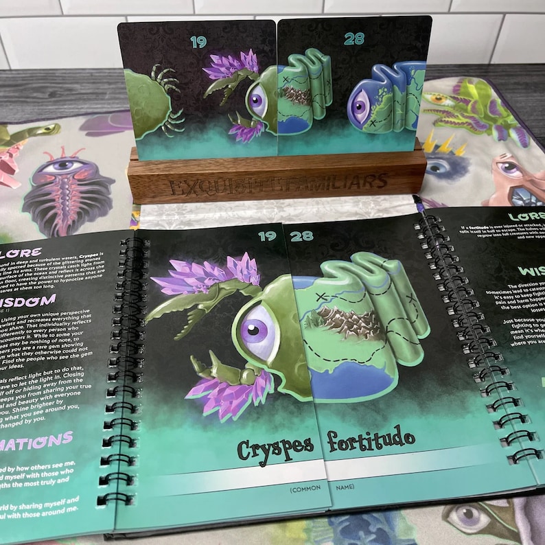 Puede incluir: Dos cartas con ilustraciones de una criatura verde y morada con un solo ojo grande. Las cartas est&aacute;n etiquetadas con los n&uacute;meros 19 y 28. Las cartas est&aacute;n colocadas sobre un soporte de madera con las palabras "Exquisite Familiars" impresas en &eacute;l. Las cartas est&aacute;n abiertas para revelar una ilustraci&oacute;n m&aacute;s grande de la criatura con el texto "Cryspes fortitudo (Nombre com&uacute;n)" impreso debajo.
