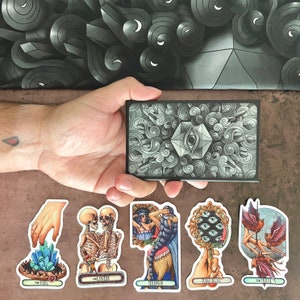 May include: A set of ten tarot cards with colorful illustrations and gold accents. The cards feature themes such as The Fool, The Lovers, Strength, The Chariot, and The Moon. Each card has a title and a corresponding image.
