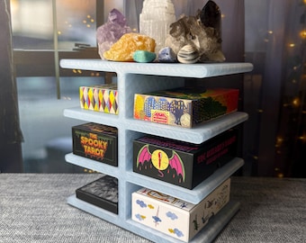 Puede incluir: Una estantería de madera azul claro que muestra cajas de cartas del tarot y cristales. La estantería contiene cajas con diseños coloridos y texto, incluyendo "The Spooky Tarot" y "Weirdos Tarot". Varios cristales en tonos morados, amarillos y transparentes están encima.