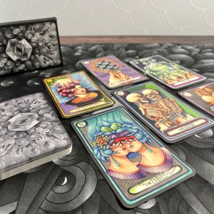 May include: A set of tarot cards with colorful illustrations and black borders. The cards feature images of a woman with three eyes, a skeleton, and a two-headed woman. The cards are laid out on a black and white patterned surface.