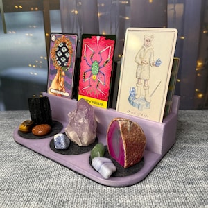 May include: A lavender-colored tarot card holder with three tarot cards and various crystals. The tarot cards feature colorful illustrations. Crystals include amethyst, agate, and other stones. The holder has a tray design with circular felt pads.