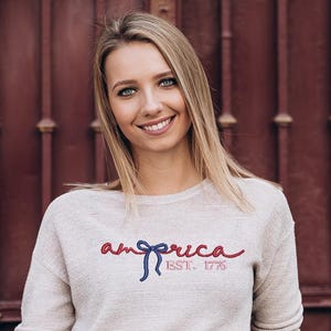 K&ouml;nnte beinhalten: Ein hellbeige Sweatshirt mit dem Wort "america" in roter Schrift, einer blauen Schleife und dem Text "EST. 1776" in Rot. Das Sweatshirt wird von einer Person mit blonden Haaren und blauen Augen getragen.