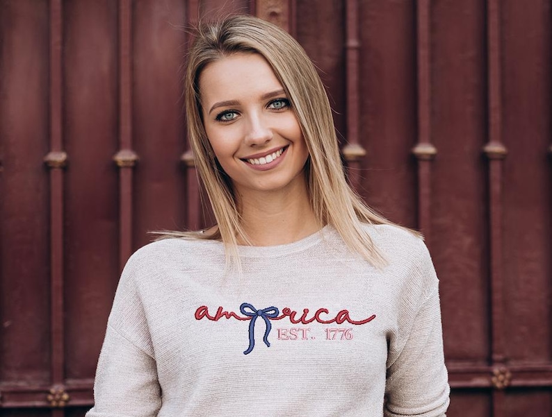 K&ouml;nnte beinhalten: Ein hellbeige Sweatshirt mit dem Wort "america" in roter Schrift, einer blauen Schleife und dem Text "EST. 1776" in Rot. Das Sweatshirt wird von einer Person mit blonden Haaren und blauen Augen getragen.