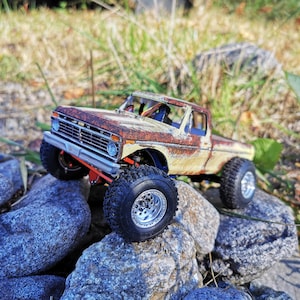 以下が含まれることがあります： さびた茶色のフードと屋根、クリーム色のボディの、ミニチュアで風化したピックアップトラック。トラックは大きな黒いオフロードタイヤを履いており、岩の上に配置されています。トラックのフロントバンパーはオレンジ色です。