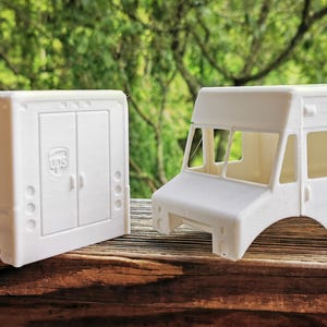 May include: Two white UPS delivery truck models. One model features a rear door design, while the other has a cab and side panel design. Both models have the UPS logo and are set against a natural background.