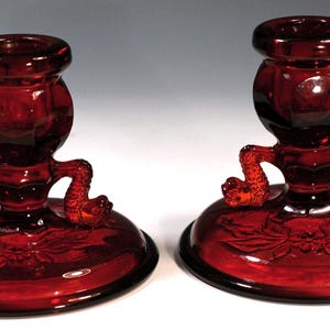 May include: A pair of ruby red glass candle holders. Each has a round base with a floral design, a central column, and a top cup for holding a candle. The candle holders have decorative, curved arms extending from the column.