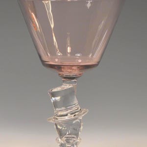 May include: A unique, clear glass goblet with a pink-tinted bowl and base. The stem features a sculpted top hat and face design. The goblet's base is round, and the bowl has a wide, conical shape.