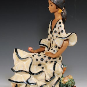 May include: A ceramic figurine of a seated figure wearing a cream-colored dress with black polka dots and ruffled edges. The figure has dark hair adorned with a small red floral accent, yellow earrings, and holds a tiny red flower in one hand.