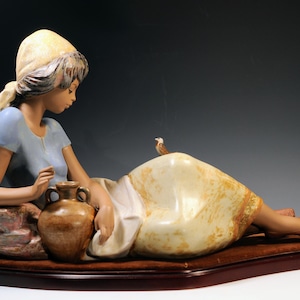 May include: A porcelain figurine of a person reclining on a textured base, wearing a light blue shirt and a cream-colored headscarf. The figure rests an arm on a brown ceramic jug, while a small bird sits on the cream-colored fabric of their skirt.