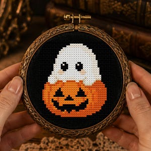 May include: A circular embroidery hoop containing a cross-stitch design on black fabric. The design features a white ghost with black eyes sitting inside an orange jack-o'-lantern pumpkin with a carved black face, all framed in a dark metal hoop.