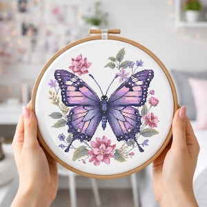 May include: A cross-stitch embroidery of a butterfly in a wooden hoop. The butterfly has purple and pink wings, surrounded by pink and purple flowers and green leaves. The embroidery is held in a wooden hoop.