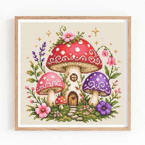 May include: A square cross-stitch embroidery in a light wood frame featuring a central large red mushroom house with white spots and decorative swirls. It is surrounded by smaller pink and purple mushrooms, green foliage, lavender sprigs, and small pink flowers on a cream background.