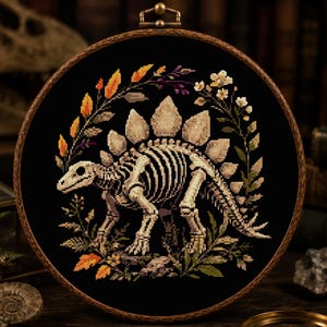 May include: A circular wooden embroidery hoop containing a cross-stitch design of a Stegosaurus skeleton on a black fabric background, surrounded by a decorative wreath of colourful autumn leaves, small white flowers, and green foliage.