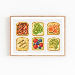 Op de afbeelding: Een ingelijst kruissteekkunstwerk met zes sneetjes toast, elk belegd met verschillende ingrediënten. De toppings zijn onder meer aardbeien en chocolade, kiwi, vijgen, bananen, gemengde bessen en avocado. Het kunstwerk staat tegen een witte achtergrond.