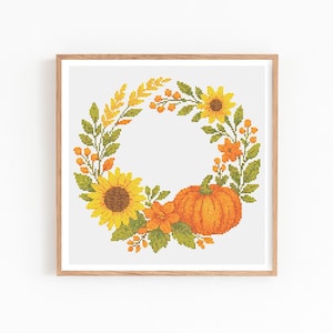 May include: A framed cross-stitch design featuring a fall wreath. The wreath includes sunflowers, an orange pumpkin, and various fall foliage in shades of yellow, orange, and green. The frame is a light brown color.