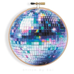 Puede incluir: Una bola de discoteca plateada con azulejos espejados, rodeada de un aro de bordado de madera. La bola refleja la luz, creando un efecto brillante.