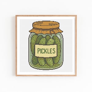May include: A framed print featuring a jar of pickles. The jar is filled with green pickles and has a yellow label that reads "PICKLES". The jar has a brown lid and is set against a white background.