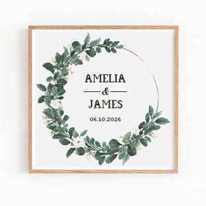 Könnte beinhalten: Gerahmter Druck mit einem floralen Kranzdesign. Der Kranz umgibt einen goldfarbenen Kreis mit den Namen "AMELIA & JAMES" und dem Datum "06.10.2026" in einer einfachen Schriftart. Der Kranz besteht aus grünen Blättern und weißen Blüten.