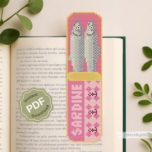 Könnte beinhalten: Handgefertigter Lesezeichen mit Kreuzstich-Design. Oben sind zwei lächelnde Fische abgebildet, darunter der Name "SARDINE" und dekorative Elemente. Das Lesezeichen ist in Rosa, Gelb und Weiß gehalten.
