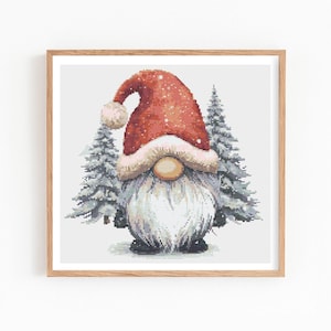 Puede incluir: Un bordado de punto de cruz enmarcado de un gnomo navideño con un gorro rojo con un pompón blanco y una larga barba blanca. Dos pinos cubiertos de nieve flanquean el gnomo. La obra de arte está sobre un fondo blanco.