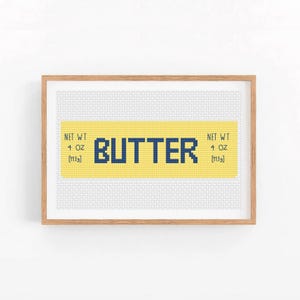 May include: A framed cross-stitch design with a yellow background and blue text that reads "BUTTER" with the words "NET WT 4 OZ [113g]" on either side of the word "BUTTER".