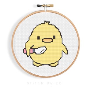 Puede incluir: Un pollito amarillo bordado a punto de cruz sobre tela blanca en un aro de bordado de madera. El pollito sostiene un cuchillo blanco en su ala derecha.