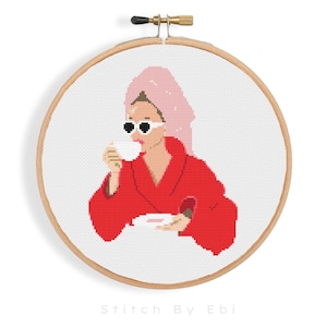 May include: A cross-stitch pattern of a woman in a red dressing gown and a pink towel on her head, drinking from a white cup. She is wearing sunglasses and holding a plate with a pink object on it.