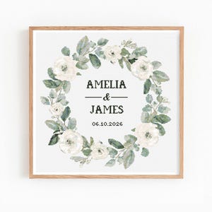 May include: A framed print featuring a floral wreath design. The wreath is composed of white roses and green leaves, surrounding the names "AMELIA & JAMES" and the date "06.10.2026" in black text. The frame is made of light-colored wood.