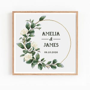 May include: A framed cross-stitch design with a floral wreath of white roses and green leaves. The design includes the names "AMELIA & JAMES" and the date "06.10.2026" within a gold-coloured circle.