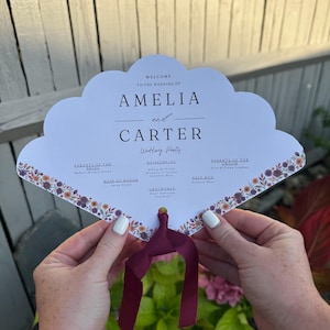 May include: A white, cloud-shaped wedding program fan with floral border and claret ribbon. The program reads "Welcome to the Wedding of Amelia and Carter" with the wedding party names listed.