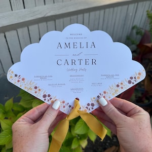 May include: A white wedding program in a cloud shape, with the names Amelia and Carter. The program includes the wedding party details and is decorated with floral accents and a yellow ribbon.