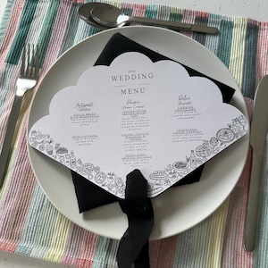 Op de afbeelding: Een trouwmenu met een geschulpte rand, met menu-items en decoratieve illustraties. De kaart ligt op een wit bord met een zwarte servet en een zwart lint. Zilverwerk en een gestreepte doek zijn ook zichtbaar.