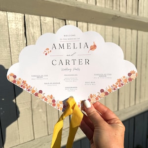 Puede incluir: Un abanico de boda blanco con borde festoneado, con los nombres "Amelia y Carter" y detalles de la fiesta de bodas. El abanico tiene detalles florales en naranja, rojo y amarillo, y se sujeta con una cinta amarilla.