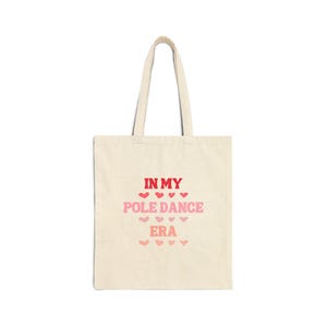 Può includere: Una borsa tote beige con il testo "In My Pole Dance Era" stampato in rosso e rosa. Il testo è circondato da una fila di cuori rosa.