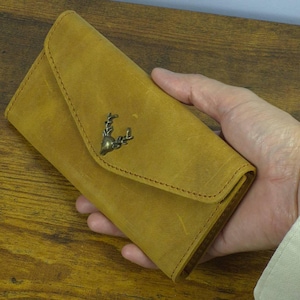 Puede incluir: Cartera de cuero color mostaza con cierre de solapa. La cartera presenta un emblema decorativo de cabeza de ciervo metálico en la parte delantera. Las costuras son visibles en los bordes. La cartera se sostiene en una mano.
