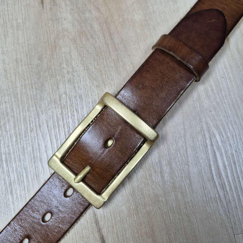 May include: Close-up of a brown leather belt with a rectangular brass buckle. The belt shows wear and has several oval holes. The buckle is a bright gold colour.
