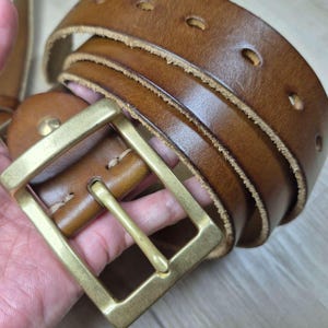May include: A brown leather belt with a brass buckle. The belt features multiple adjustment holes and a classic design. The leather has a worn appearance, giving it a vintage look. The belt is approximately 100 cm in length.