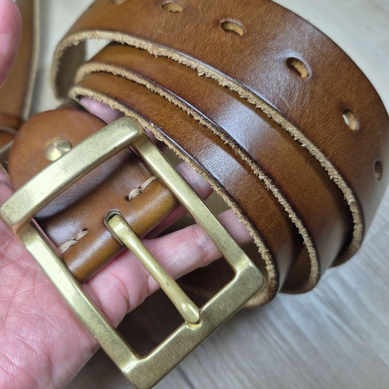 May include: A brown leather belt with a brass buckle. The belt features multiple adjustment holes and a classic design. The leather has a worn appearance, giving it a vintage look. The belt is approximately 100 cm in length.