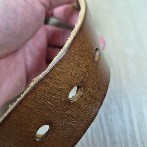May include: Close-up of a brown leather belt with several evenly spaced holes. The belt has a textured surface and a lighter-coloured edge. The leather appears worn, suggesting a vintage or rustic style.