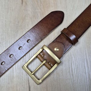 May include: A brown leather belt with a brass buckle. The belt features a rectangular buckle and multiple adjustment holes. The leather has a rich, warm colour. The belt is designed for everyday use and is suitable for trousers or jeans.