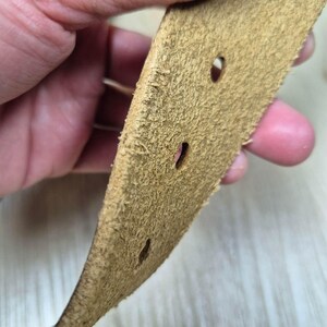 May include: Close-up of a tan leather belt with three oval holes. The belt has a textured, suede-like appearance and is held by a hand. The edges of the belt are frayed.