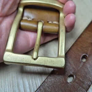 May include: Close-up of a brown leather belt with a brass buckle. The buckle is rectangular with a single prong. The leather belt has two holes visible. The belt and buckle are held by a person.