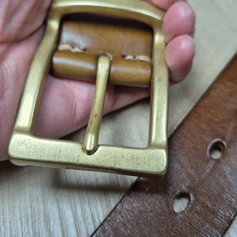 May include: Close-up of a brown leather belt with a brass buckle. The buckle is rectangular with a single prong. The leather belt has two holes visible. The belt and buckle are held by a person.