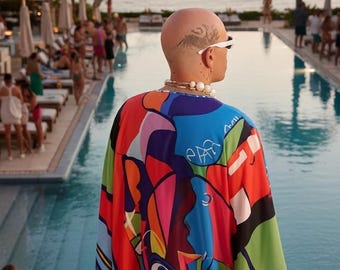 May include: A person wearing a colorful, abstract patterned cape with blue, red, green, and yellow hues. The person is wearing white sunglasses and a pearl necklace. The background includes a pool, ocean, and palm trees.