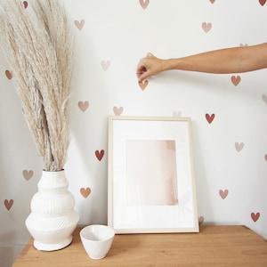 May include: A white vase with a textured surface filled with dried pampas grass sits on a wooden table next to a white picture frame and a white bowl. The wall behind the vase is decorated with a pattern of small, pink hearts.
