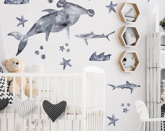 May include: A nursery wall decorated with watercolor shark and ocean-themed decals in shades of grey and blue. The design includes a hammerhead shark, various shark silhouettes, stars, and bubbles. A white cot and decorative shelves are also visible.