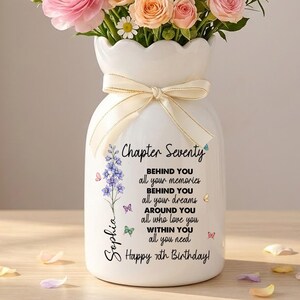May include: A white ceramic vase with a scalloped rim and a cream-colored ribbon tied around the neck. The vase features text that reads: Chapter Seventy, BEHIND YOU all your memories, BEHIND YOU all your dreams, AROUND YOU all who love you, WITHIN YOU all you need, Happy 70th Birthday, Sophia. It is filled with a bouquet of pink, peach, and white flowers including ranunculus and daisies.